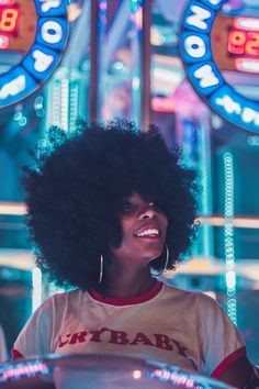 a woman with an afro smiles in front of neon signs and lights at the fairground