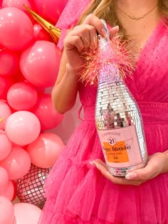 a woman in a pink dress holding a bottle of booze next to balloons and streamers