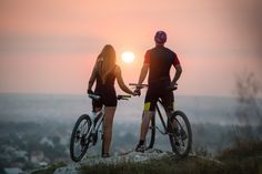 two people standing on top of a hill with their bikes looking at the sun setting