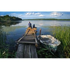 two people are sitting on a dock with a boat in the water and grass around them