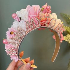 a hand holding a tiara with pink flowers and pearls on the front, in front of a mirror