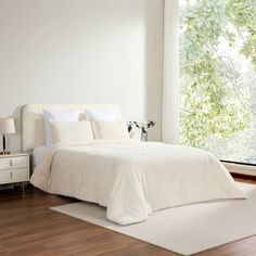 a white bed sitting on top of a wooden floor next to a large window in a bedroom