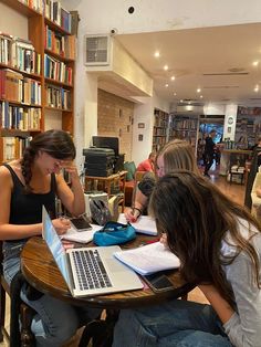 three women sitting at a table working on laptops in a library with bookshelves behind them