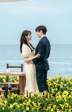 two people standing next to each other in front of flowers and an umbrella near the ocean