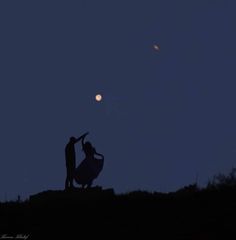 two people standing on top of a hill at night with the moon in the background