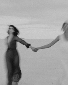 two women holding hands while walking on the beach with their arms around each other, in black and white