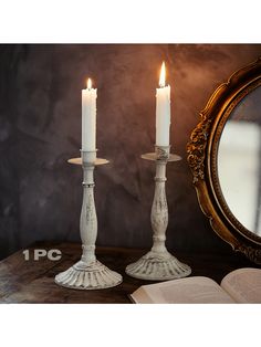 two white candles sitting on top of a table next to an open book and mirror