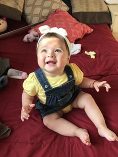 a baby sitting on a bed wearing overalls and a bow in her hair, smiling at the camera