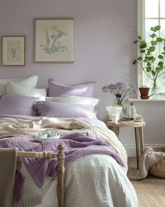 a bed with purple sheets and pillows in a bedroom next to a window filled with potted plants