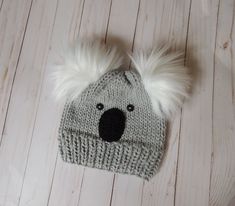 a knitted koala hat with white fur pom - poms sits on a wooden floor