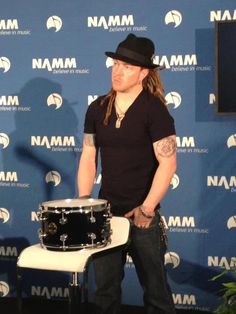 a man with long hair and a hat standing in front of a drum on a chair