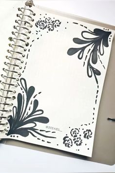 an open spiral notebook with black and white designs on it, next to a pen