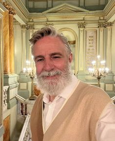 a man with grey hair and beard standing in front of a staircase wearing a vest