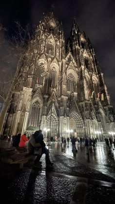 people sitting on benches in front of a large cathedral at night with the lights on