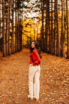 a woman standing in the middle of a forest with leaves on the ground and trees behind her