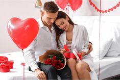 a man and woman sitting on a bed with red balloons in the shape of hearts