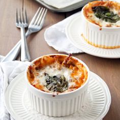 two white dishes filled with lasagna and spinach on top of a wooden table