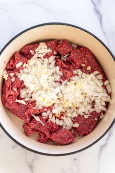 a bowl filled with meat and onions on top of a white marble countertop next to a wooden spoon