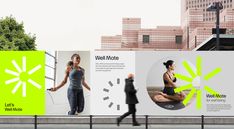 two billboards on the side of a road with people walking past them and buildings in the background