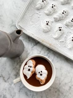 two cookies in the shape of ghost sitting in a cup of hot chocolate next to a cookie sheet