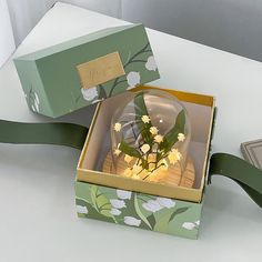 an open box with flowers inside on a white table next to a green ribbon and card