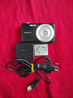 a digital camera sitting on top of a red blanket next to a charger and power cord