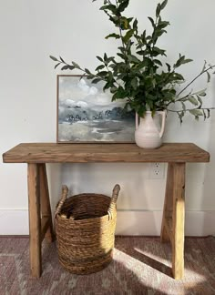 a wooden table topped with a basket next to a vase filled with green leaves and a painting on the wall