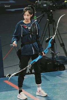 a woman holding a bow and arrow on top of a blue floor in front of a camera