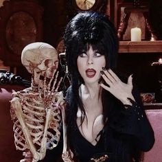 a woman with black hair and makeup holding her hands up to her face next to a skeleton