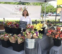 a woman standing in front of flowers at a farmers market with signs on the tables