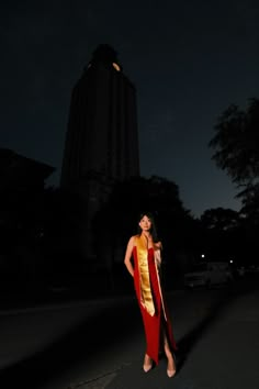 a woman in a red and gold dress standing on the street at night with her hands behind her back