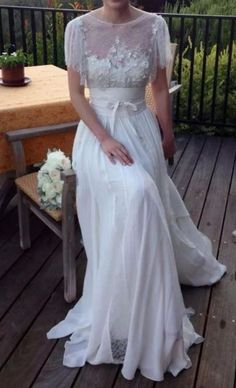 a woman in a white dress sitting on a wooden deck next to a table with flowers