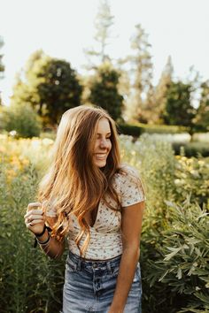 a woman with long hair standing in front of some bushes and flowers smiling at the camera