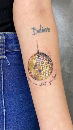 a woman's arm with a tattoo on it that says believe and an image of a disco ball