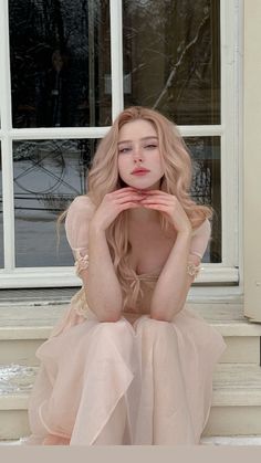 a woman with long blonde hair sitting in front of a window wearing a pink dress