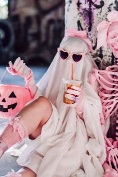 a woman dressed as a ghost sitting next to a halloween tree with a drink in her hand