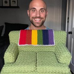 a man is smiling while sitting in a green chair with a rainbow crochet pillow on it