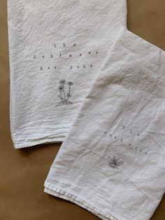 two white tea towels with words on them sitting on a brown tablecloth covered surface