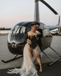 a man and woman standing in front of a helicopter on the tarmac with their arms around each other