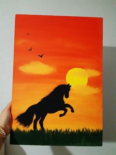 a person holding up a painting of a horse in front of an orange and red sky