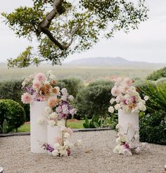 three white vases filled with flowers sitting on top of a gravel covered ground next to trees