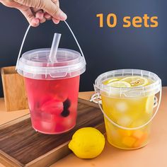 two plastic cups with lemons and drinks in them on a wooden tray next to a hand holding a straw