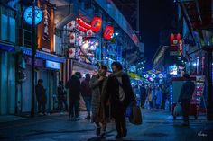 two people are walking down the street at night with neon signs on buildings in the background