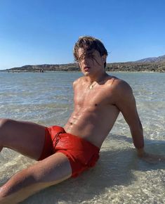 a shirtless man sitting on the beach in shallow water with his legs spread out