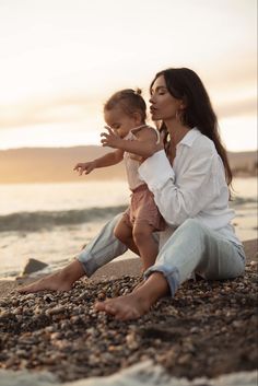 a woman holding a baby sitting on top of a sandy beach next to the ocean