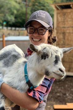 a woman holding a goat in her arms and smiling at the camera while wearing a baseball cap