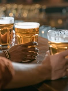 two hands holding glasses of beer on a table with other people in the background at a bar