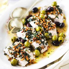 a white plate topped with fish covered in olives and capers next to a spoon