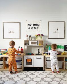 two toddlers playing in a play kitchen with toys on the floor and wall behind them