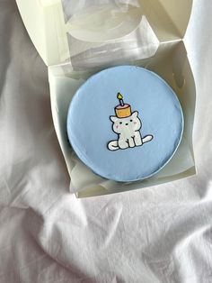 a blue cake with a cat on it in a white box, sitting on a bed